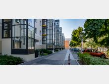 Moderne Wohngebäude mit verglasten Aufzugsschächten an der Fassade entlang eines gepflasterten Gehwegs, flankiert von Grünflächen, geparkten Autos und Bäumen. Im Hintergrund ist ein orangefarbenes Gebäude zu sehen.