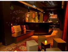 Gemütliche Bar mit dunklem Interieur, Holzboden und gedämpfter Beleuchtung. Im Vordergrund befinden sich gepolsterte Hocker und niedrige Tische mit kleinen Kerzen, im Hintergrund eine geschwungene Theke vor einer mit floralen Mustern bemalten Wand.