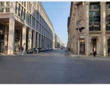 Straßenszene in Berlin mit einem breiten Gehweg und mehrstöckigen Gebäuden aus heller Steinfassade links und rechts. Links steht ein modernes Gebäude mit Arkadengang, rechts ein historisch verziertes Gebäude, daneben Autos und einige Fußgänger sowie Radfahrer.