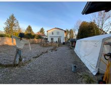 Ein einfacher Garten mit einem Scarecrow aus Alltagskleidung im Vordergrund, einem weißen Einfamilienhaus im Hintergrund und einer Seitenplane eines Gewächshauses rechts. Der Garten ist teilweise mit Kieswegen und Holzzäunen eingefasst, mit Bäumen und Sträuchern rundherum.