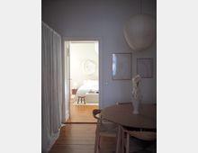Minimalist apartment dining area with a round wooden table and chairs, sheer floor-to-ceiling curtains, framed wall art, and a large paper pendant light. An open doorway reveals a bright bedroom with a white bed, bedside table and lamp, and light wood floors throughout.