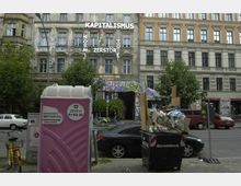 Mehrstöckiges Altbaugebäude mit verwitterter Fassade, bemalt mit politischen Aussagen und Graffiti; im Vordergrund eine rosafarbene mobile Toilettenkabine und ein überfüllter Abfallcontainer auf einem Bürgersteig. Auf der Straße parken Autos, umgeben von Bäumen, und dahinter befindet sich weiteres leicht renoviertes Altbauwerk.