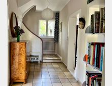 Ein heller Flur mit hellbeigen Wänden und beige-braunen Fliesen, einer geschwungenen Treppe mit weißem und dunklem Holzgeländer, einem runden weißen Lampenschirm und einem Fenster mit Heizung darunter. Links steht eine Holzkommode mit runder Spiegeldeko darüber, rechts ein Bücherregal und eine Wandmaske.