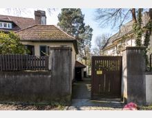 Einfamilienhaus mit verputzter Fassade und rotem Ziegeldach, umgeben von einer Betonmauer und einem Holztor mit einem Schild "Einfahrt freihalten". Im Hintergrund sind Bäume und ein weiteres Haus sichtbar.
