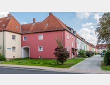 Reihenhäuser in einer Wohnsiedlung mit hellen Fassaden in Rot, Beige und Pastelltönen. Die Gebäude haben ziegelrote Dächer, kleine Vorgärten und angrenzende Gehwege, daneben eine gepflasterte Straße.