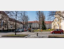 Wohnquartier mit gelb gestrichenen Mehrfamilienhäusern, roten Ziegeldächern und großen Fenstern. Im Vordergrund verlaufen eine Straße mit geparkten Autos und Motorrädern sowie ein kleiner begrünt angelegter Platz mit kahlen Bäumen.