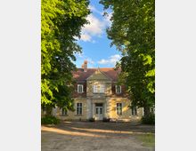 Historisches Gebäude mit gelber Fassade und rotem Ziegeldach, flankiert von hohen Bäumen. Der Eingangsbereich verfügt über eine Symmetrie mit einem Dreiecksgiebel und großen Fenstern.