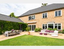 Backstein-Einfamilienhaus mit gepflegtem Garten und großer Terrasse. Links befindet sich ein mit Efeu bewachsener Anbau mit einer langen Esstischgruppe, rechts eine Sitzecke mit Sofa, lila Kissen und Hockern auf dem Terrassenboden.