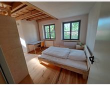 Ein schlicht eingerichtetes Schlafzimmer mit Holzboden, einem Bett aus hellem Holz, einem kleinen Schreibtisch mit Stuhl und zwei großen Fenstern, die Blick auf grüne Bäume bieten. Die Holzdecke zeigt sichtbare Balken, und die Wände sind weiß gestrichen.