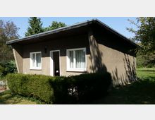 Ein kleiner, einfacher Bungalow mit flachem Dach und brauner Fassade, zwei Fenstern mit weißen Rahmen und einer offenen Tür. Das Gebäude ist von einem gepflegten Garten mit Büschen und Rasen umgeben, im Hintergrund stehen Bäume.