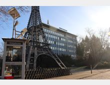 Nachbildung des Eiffelturms aus Metall, daneben eine umgebaute Telefonzelle, die als Bücherschrank dient. Im Hintergrund ein modernes Bürogebäude mit der Aufschrift "Centre Français", umgeben von Bäumen.