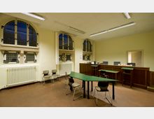 Innenraum eines Verwaltungsraums mit Holztheke, mehreren grünen Schreibtischen, dazugehörigen Stühlen und hohen, bogenförmigen Fenstern mit Gitterdetails. Der Raum ist mit cremefarbenen Wänden, einer einfachen Deckenbeleuchtung und Heizkörpern ausgestattet.