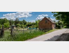 Ein ländlicher Weg mit einer alten Backsteinscheune auf der rechten Seite, umgeben von einem Holzzaun und einer Wiese. Im Hintergrund befinden sich Gärten mit kleinen Hütten, Bäumen und Sträuchern, unter einem bewölkten blauen Himmel.