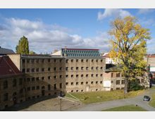 Backsteingebäude eines historischen Komplexes mit mehreren Fenstern und einer Glasdachkonstruktion im oberen Bereich, umgeben von einem gepflasterten Hof und Rasenflächen mit Herbstlaub. Im Vordergrund steht ein großer Baum, und der Himmel ist teilweise bewölkt.