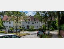 Reihenhäuser mit heller Fassade und roten Ziegeldächern, umgeben von Bäumen und grünem Bewuchs. Im Vordergrund führt eine gepflasterte Straße mit parkenden Autos durch eine ruhige Wohngegend.