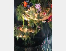 Eine dekorative Deckeninstallation mit goldenen Lampenschirmen, umgeben von üppigem Grün und bunten künstlichen Blumen. Die Wände sind dunkel und mit einem floralen Muster verziert.