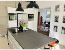 Moderne Küche mit Kücheninsel aus dunklem Stein, eingebautem Kochfeld und Hockern. Im Hintergrund ein Essbereich mit Holzboden, bunten Stühlen und großem Fenster, sowie eine Wand mit Schwarz-Weiß-Fotografien.