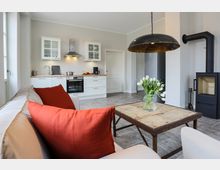 Offene Wohnküche mit weißen Einbaumöbeln, Herd und Dunstabzugshaube, daneben ein moderner Kaminofen. Im Vordergrund stehen ein Sofa mit roten Kissen und ein rustikaler Holztisch mit einer Vase weißer Tulpen.