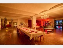 Großer, warm beleuchteter Veranstaltungsraum mit Holzboden, niedriger Decke und roten Vorhängen, in der Mitte ein lang gedeckter Esstisch mit Kerzenleuchtern. Rechts steht ein schwarzer Flügel unter einem Kronleuchter, dahinter große Fenster und eine offene Tür mit Blick auf die nächtliche Straße.