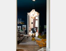 Wohnzimmer mit modernem Design und blauen Wänden, ausgestattet mit einem grauen Sofa, gelben und weißen Stühlen sowie einem runden Glastisch. An der Decke hängt eine geometrische Lampe, und durch eine offene Tür ist ein angrenzender Raum mit Vorhängen und großen Fenstern sichtbar.