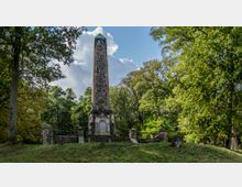 Ein steinerner Obelisk steht auf einer kleinen Anhöhe inmitten eines Parks umgeben von dichtem Grün. Das Denkmal ist von einer niedrigen Steinmauer eingerahmt, darunter eine Gedenktafel, während im Hintergrund Bäume und Wolken den Himmel bedecken.
