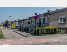 Straßenszene in einem Wohngebiet mit einfachen einstöckigen Häusern, Vorgärten und Gehwegen. Verkehrsschilder zeigen eine Tempo-30-Zone und ein Halteverbot, und ein rot markierter Fahrradweg kreuzt die Straße, während zwei Radfahrer auf dem Gehweg fahren.