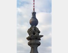 Eine Steinverzierung in Form abstrakter Flammen im Vordergrund, mit dem Berliner Fernsehturm und einem bewölkten Himmel im Hintergrund. Der Fernsehturm hat seine markante Kugel mit Antenne und roten Segmenten.