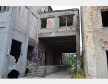 Verfallenes Fabrikgebäude mit verwitterten Beton- und Ziegelwänden, teils eingestürzten Fenstern und bemalter Fassade. Ein Torbogen führt in einen überwucherten Innenbereich mit Pflanzen.