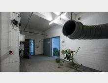 Industrie- oder Technikraum mit weiß gestrichenen Ziegelwänden, einer offenen Stahltür und einem großen metallischen Lüftungsrohr. Im Vordergrund steht ein hand- oder fußbetriebenes Gerät mit Sitz, vermutlich für die Luftzirkulation.