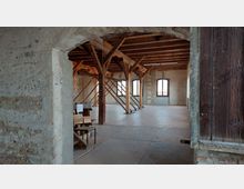 Ein leerer Raum in einem historischen Gebäude mit freiliegendem Mauerwerk, Holzbalken und großen Fensteröffnungen. Im Vordergrund ist ein steinerner Türrahmen und einige Stühle sichtbar.