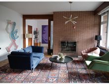 Wohnzimmer mit modernem und retro-inspiriertem Design; eine Backsteinwand mit integriertem Kamin, große Fenster rechts, ein blauer Samtsofa, ein roter und ein grüner Sessel sowie ein runder Couchtisch auf einem gemusterten Teppich. Wanddekoration und Kunstobjekte ergänzen den Raum.