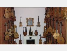 Raum mit weißen Wänden und einem dekorativen Kamin, umgeben von zahlreichen historischen Zupfinstrumenten wie Gitarren und Lauten, die an den Wänden aufgehängt sind. Über dem Kamin hängt ein goldgerahmter Spiegel, darunter stehen verzierte Harfen.