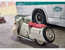 Oldtimer-Motorroller mit Seitenwagen und roten Sitzpolstern steht auf einer betonierten Fläche unter einem überdachten Bereich. Im Hintergrund ist die Seitenwand und ein großes Rad eines Busses zu sehen, dahinter eine Grünfläche.