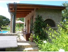 Kleines Gebäude aus Backstein mit Rundbogenfenster und hölzernem Vordach, umgeben von üppigem Grün und blühenden Pflanzen. Im Hintergrund sind ein Pool, Gartenmöbel und ein Sonnenschirm auf einer Terrasse sichtbar.