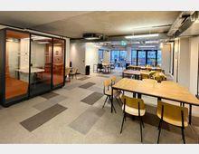 Ein moderner Coworking-Bereich mit Holztischen, schlichten Holzstühlen und einstellbaren Hängelampen auf Teppichboden. Links ist eine schallisolierte Meeting-Kabine mit Glastüren und orangefarbenen Polstern, rechts führen Fensterfronten mit Blick auf die Straße viel Tageslicht in den Raum.