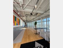Open-plan modern office or studio with wood floors, an exposed white ceiling with pipes and track lighting, and floor-to-ceiling windows along one side overlooking a city. The space includes a long counter with colorful wall art, a potted plant, and scattered tables and lounge seating.