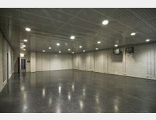 Leerer Raum mit grauen Betonwänden, glänzendem dunklen Boden und einer Decke mit eingebauten Deckenleuchten. An den Wänden hängen Kabel und zwei ältere Fernseher, links ist eine Tür zu sehen.