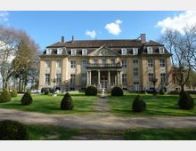 Historisches Herrenhaus mit symmetrischer Fassade, hohen Fenstern und einem zentralen Säulenvorbau. Das Gebäude befindet sich in einer gepflegten Parkanlage mit Rasen, Gehwegen und dekorativen Buchsbaumbüschen.