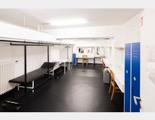 Ein heller Umkleideraum mit weißen Wänden und einer schwarzen Bodenfläche. Der Raum ist mit Schminktischen, Holzstühlen, blauen Schließfächern, einem Heizkörper und einer Kleiderstange ausgestattet; Spiegel sind über die Tische angebracht.