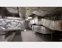 Technikraum mit umfangreichen Belüftungsanlagen, bestehend aus großen, silberfarbenen und isolierten Luftkanälen, die an der Decke und den Wänden verlaufen. Die Beleuchtung erfolgt über an der Decke montierte Leuchtstofflampen.