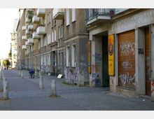 Straßenansicht mit einer Reihe von mehrstöckigen Wohngebäuden, deren Fassade teilweise abgenutzt und mit Graffiti bedeckt ist. Die Gebäude haben kleinere Balkone mit Blumenkästen, daneben stehen Betonpoller entlang eines gepflasterten Gehwegs, und ein Fahrrad ist an einem Poller angelehnt.