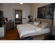 Ein Schlafzimmer mit einem großen, gepolsterten Bett und weißer Bettwäsche, flankiert von Nachttischen mit Lampen. Im Raum befinden sich ein seitlicher Schrank, ein Spiegel, und eine dekorative Wand mit einem großformatigen Schwarz-Weiß-Bild einer Stadtansicht. Im Hintergrund ist eine geöffnete Tür zu einem weiteren Raum zu erkennen.