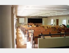 Ein Hörsaal mit ansteigenden Sitzreihen aus Holz und integrierten Tischen, Blickrichtung zur Vorderseite mit einer großen Tafel und einer Projektionsfläche. Der Raum ist gut beleuchtet durch rechteckige Deckenlampen und Tageslicht, das durch mehrere Fenster an der rechten Wand einfällt.