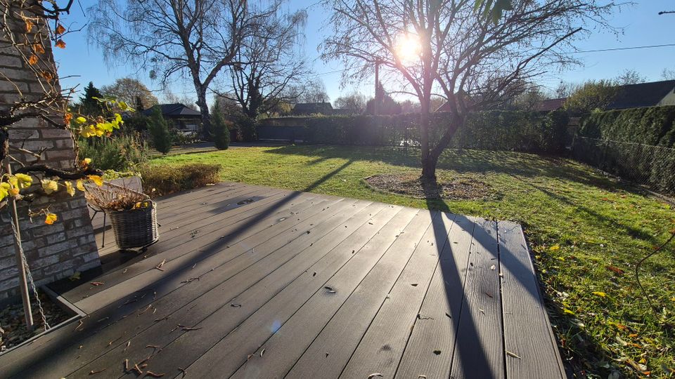 Sonniger Garten mit einer Holzterrasse im Vordergrund, umgeben von einer Rasenfläche mit einem großen Baum, kahlen Ästen und vereinzeltem Laub. Im Hintergrund sind Bäume, Sträucher und ein Zaun zu sehen.