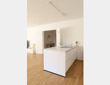 Moderne, minimalistisch gestaltete Küche mit einer weißen Kochinsel und heller Holzoptik auf dem Boden, im Hintergrund ein geöffneter Türdurchgang zu einem weiteren Raum. Wanddekor und neutrale Farben prägen die Umgebung.