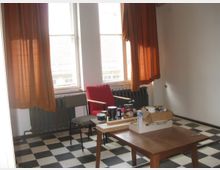 Ein einfacher Raum mit zwei Fenstern, durch die Licht fällt, braunen Vorhängen und einem schwarz-weißen Schachbrettboden. Im Raum stehen ein Holztisch mit verschiedenen Gegenständen, zwei Stühle und ein Heizkörper unter den Fenstern.