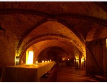 Historischer Gewölbekeller mit Ziegelmauerwerk und mehreren Rundbögen, stimmungsvoll beleuchtet durch Kerzen und warme Lichtquellen. Ein langer, weiß gedeckter Tisch dominiert den Raum, der rustikal eingerichtet ist.