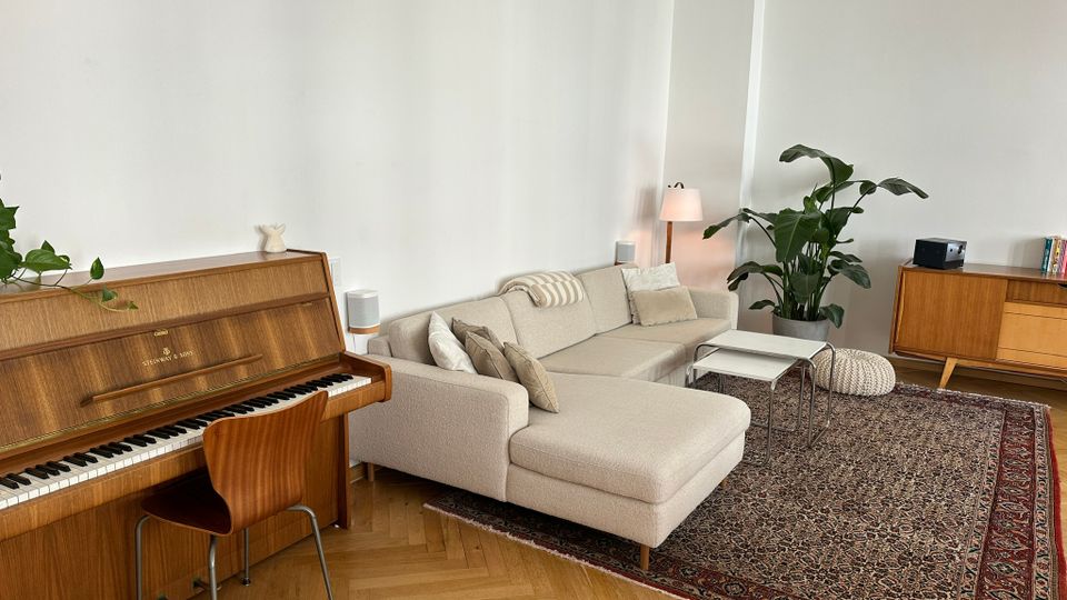 Helles Wohnzimmer mit weißer Wand, Parkettboden im Fischgrätmuster und großem gemustertem Teppich. Im Raum stehen ein beiges Ecksofa mit Couchtisch, ein Klavier mit Stuhl sowie ein Sideboard, eine Stehlampe und große Zimmerpflanzen.