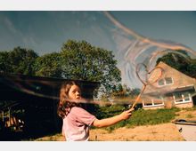 Ein Kind spielt draußen mit einer großen Seifenblasenstange, während im Hintergrund ein Haus mit Satteldach und eine grüne, ländliche Umgebung mit Bäumen zu sehen sind. Eine Scheune oder Überdachung ist ebenfalls links im Bild sichtbar.