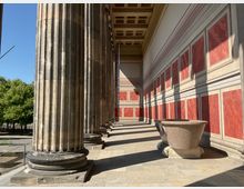 Außenansicht eines klassischen Säulengangs mit massiven, geriffelten Steinsäulen und einer rot-weiß gestrichenen Wand mit rechteckigen Vertiefungen. Im Vordergrund steht eine große steinerne Wanne auf einem Sockel, im Hintergrund sind Bäume und blauer Himmel sichtbar.